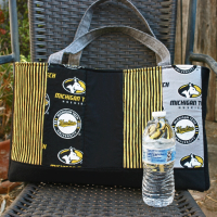 Front view. Vertical strips of MTU and coordinating fabrics. Grey straps. Water bottle (not included) to show size of bag.