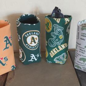 Photo of 4 different upright cylindrical upright eyeglasses holders: Gold with green As with a red heart instead of an apostrophe; green with circular A's logo; green with all over scattered A's logos, elephants and the word Oakland; and white with various A's words and sayings.